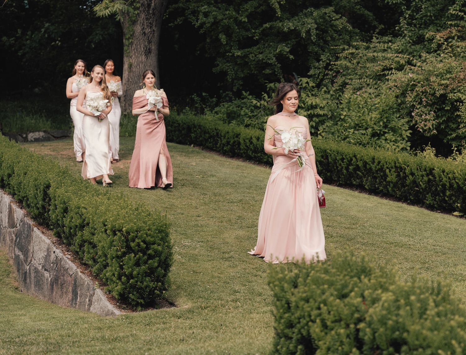 Bridesmaids in pink dresses walk through a lush garden carrying white bouquets at an outdoor wedding ceremony.
