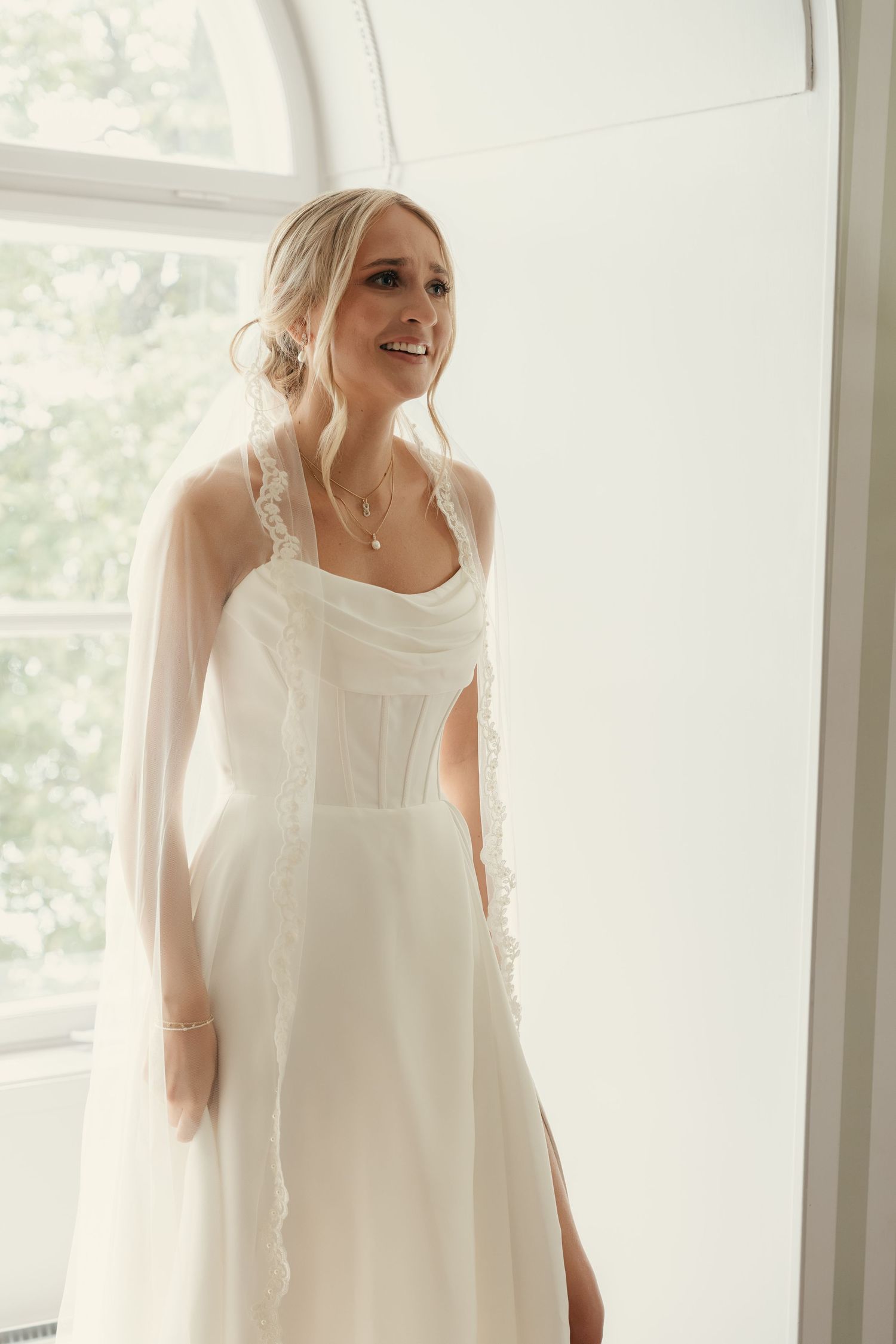 Smiling bride in elegant white corset wedding dress with sheer sleeves standing by a bright window.