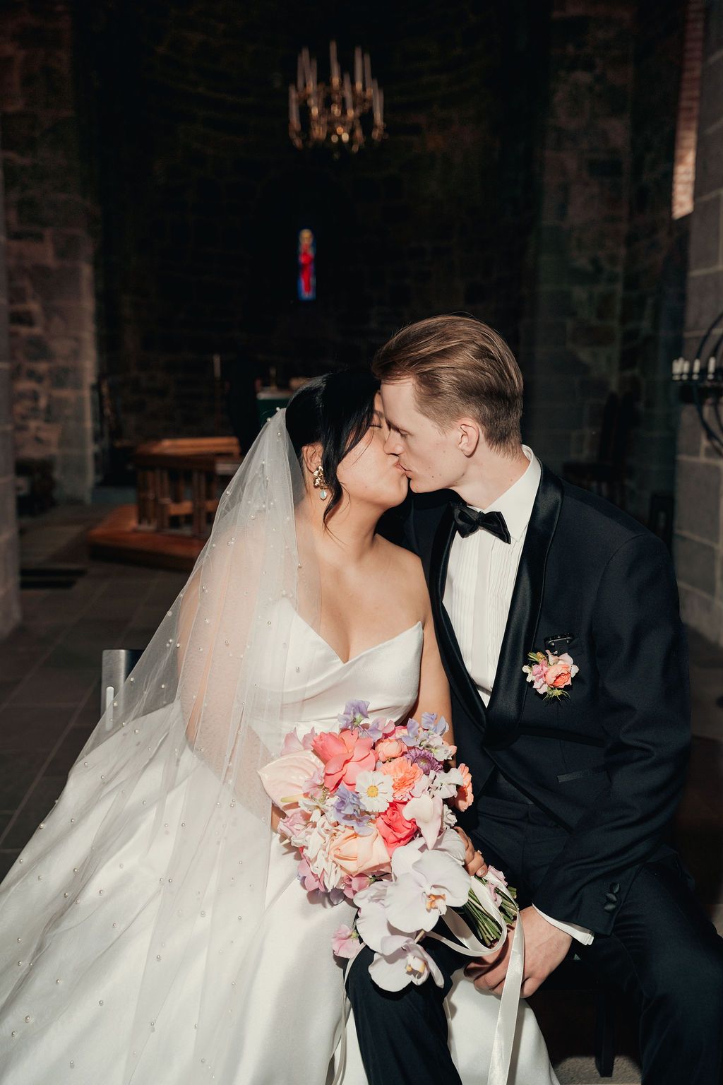 Romantic wedding couple in formal attire share an intimate moment against a rustic brick wall backdrop.