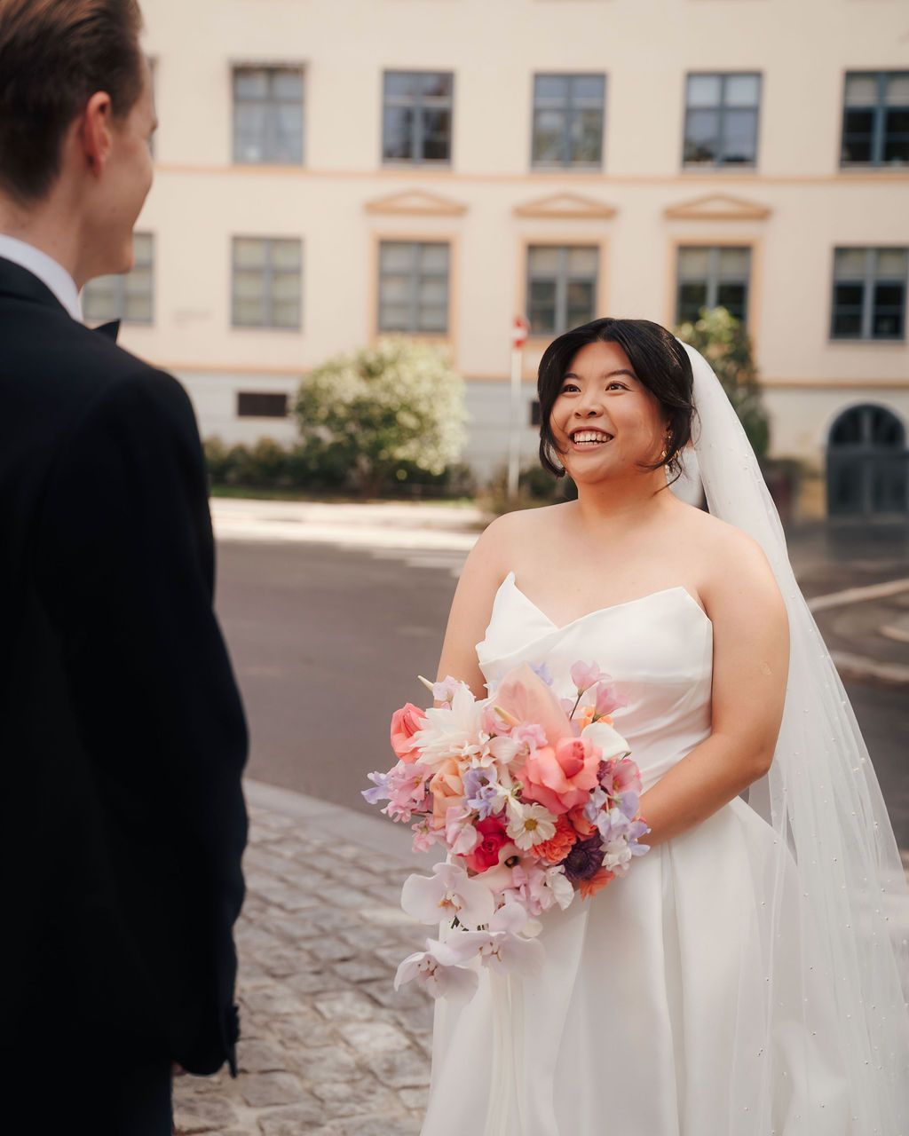 Bride in white wedding dress holds pink and coral floral bouquet while sharing a joyful moment outdoors.