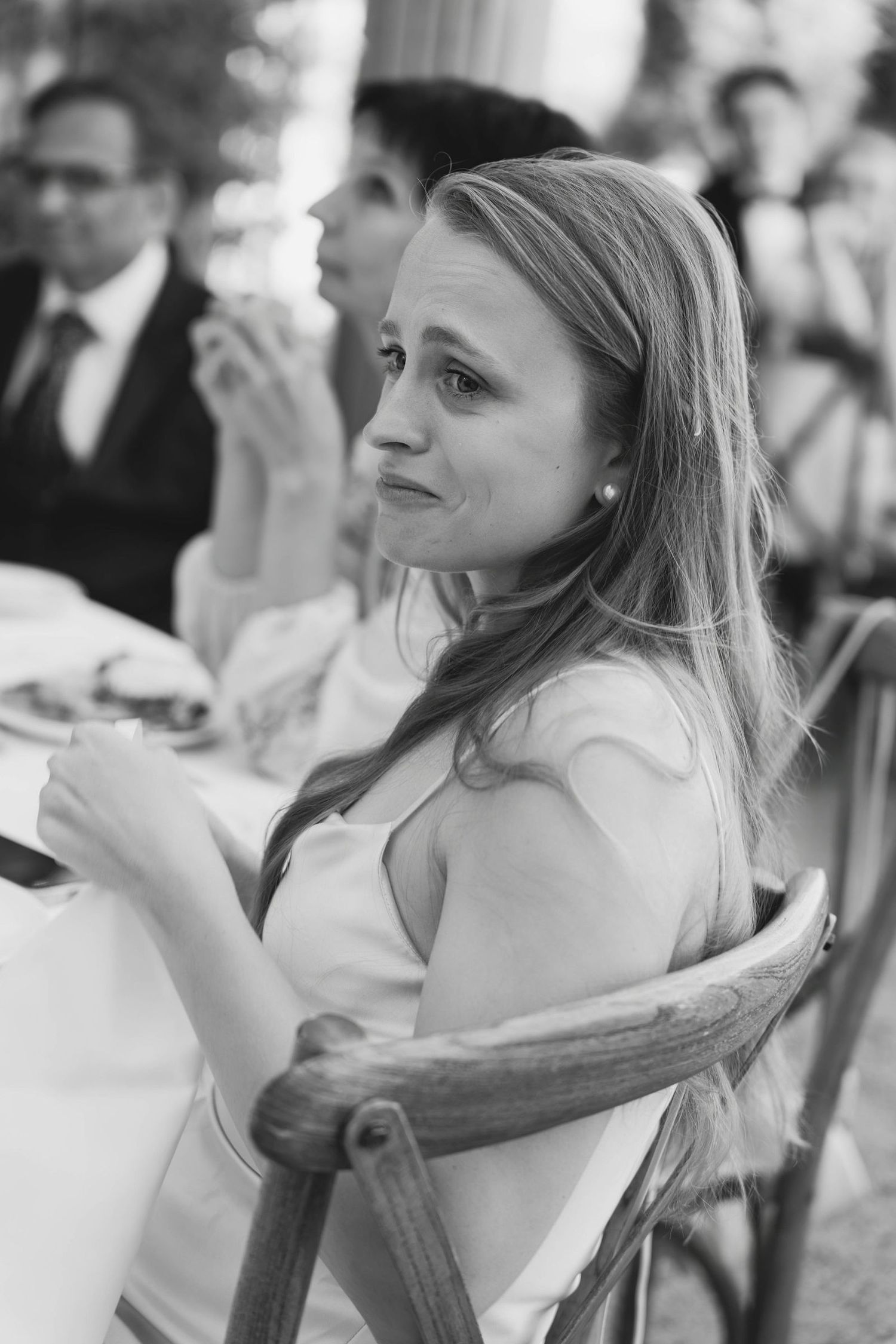 Black and white photo of a woman with long hair sitting in a wooden chair at an outdoor event.