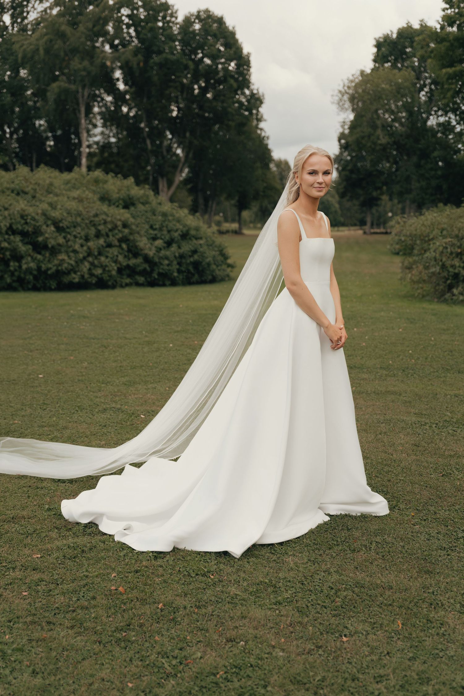 Bride in an elegant white satin gown with a long flowing veil standing on a green lawn surrounded by trees.