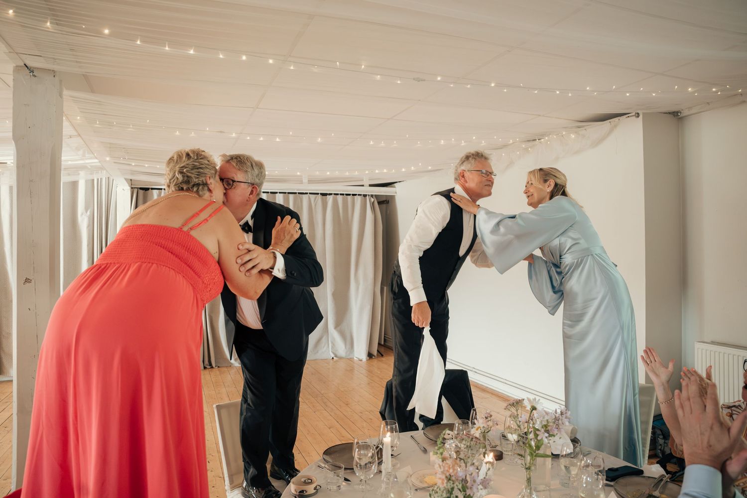 Wedding guests in formal attire greet and hug each other beside a reception table decorated with flowers.