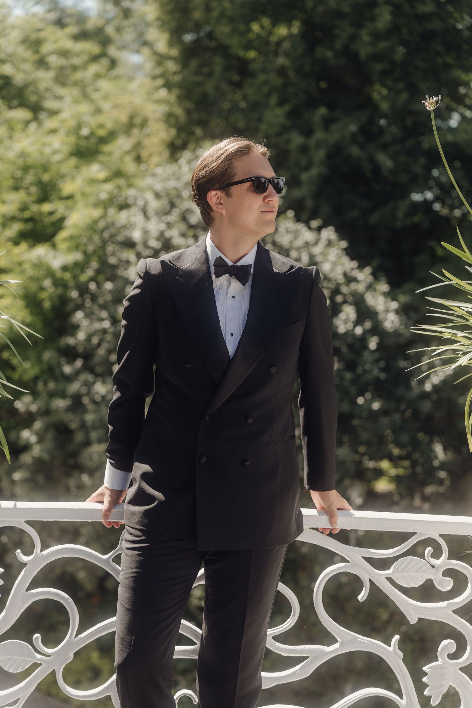 Man in a black double-breasted tuxedo, bow tie and sunglasses leaning on an ornate white railing with green foliage behind him.