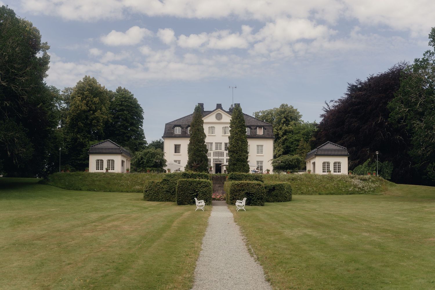 Symmetrical manor house with two flanking pavilions at the end of a gravel path through a manicured green lawn framed by tall trees.
