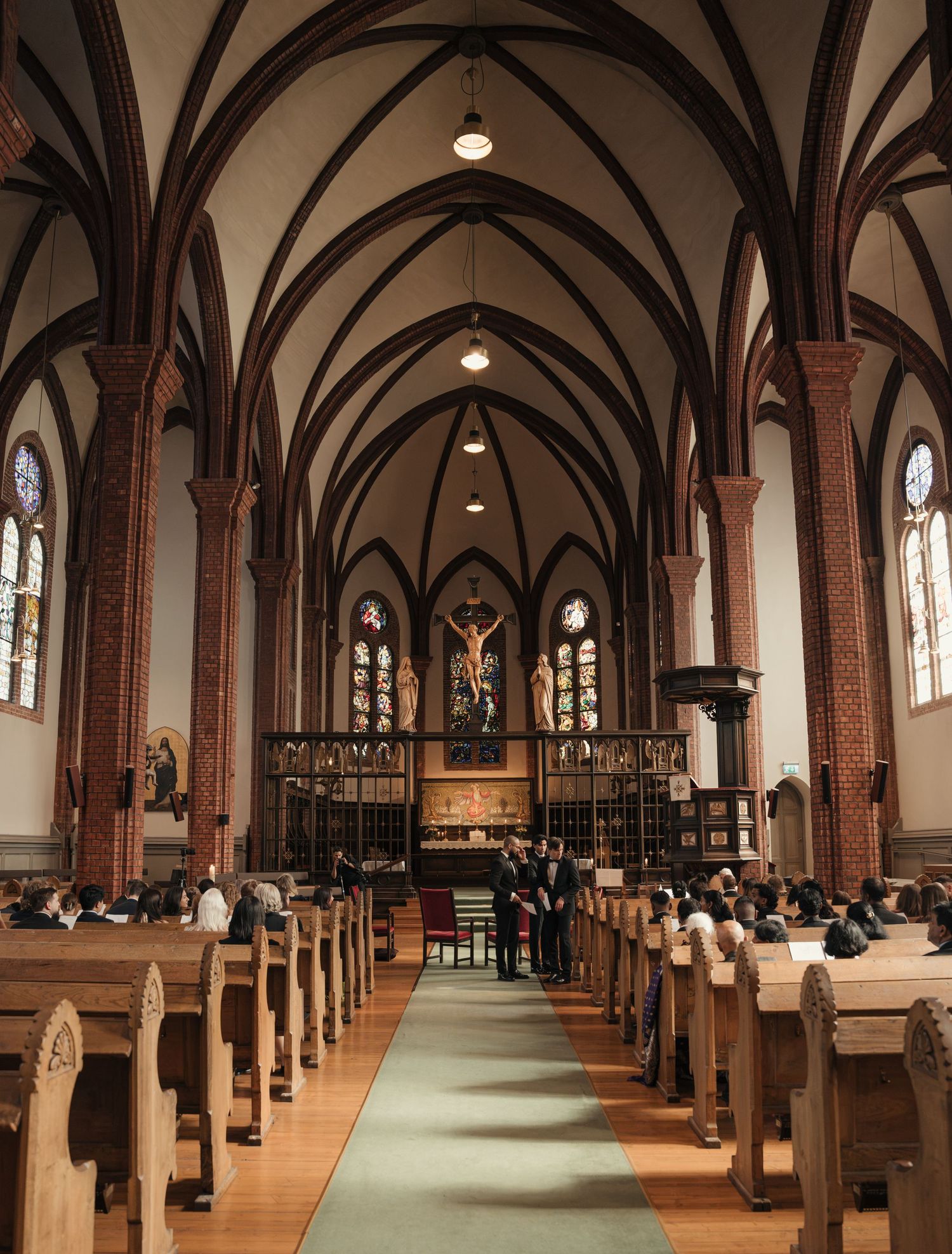 Wedding guests sit in wooden pews inside a Gothic church with red brick columns, pointed arches and a crucifix above the altar.