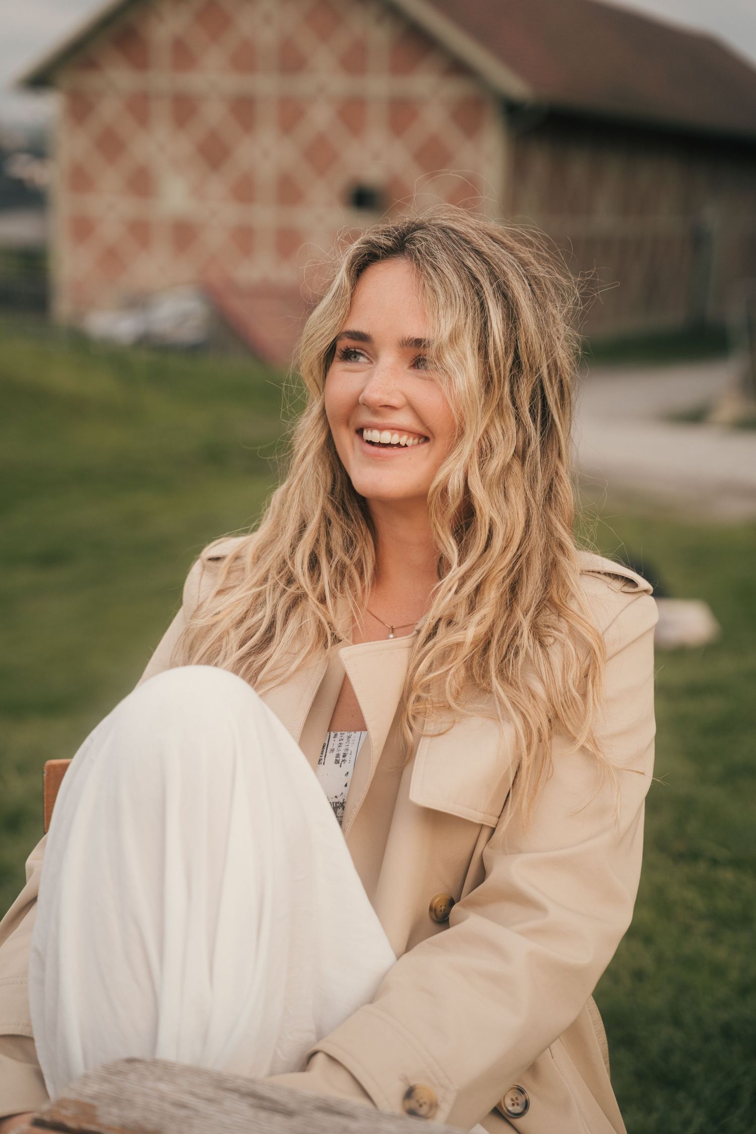 A person in a beige coat and white dress sits on grass during an outdoor photoshoot.