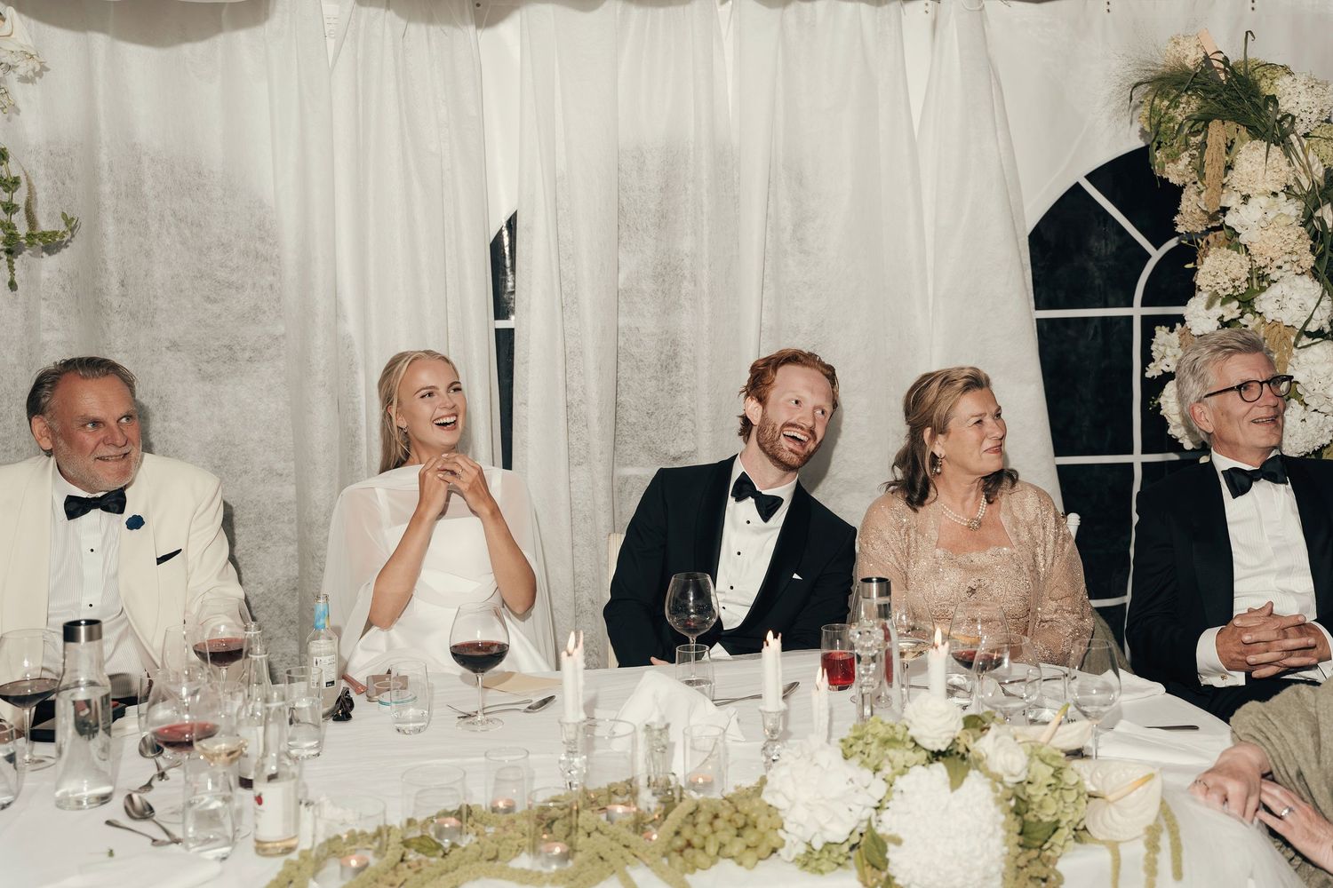 Smiling wedding guests in formal wear sit at a elegantly set reception table decorated with white flowers, candles and wine glasses.