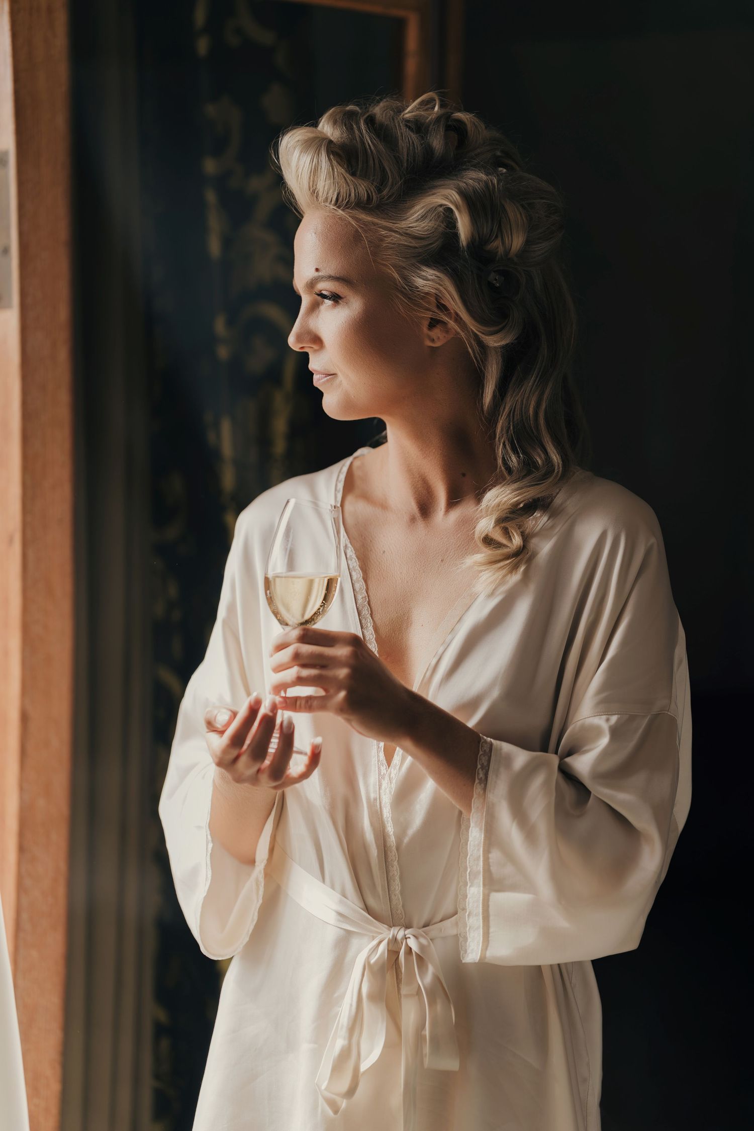 Woman in a cream satin robe with an elegant updo holds a glass of champagne while gazing out a window in soft natural light.