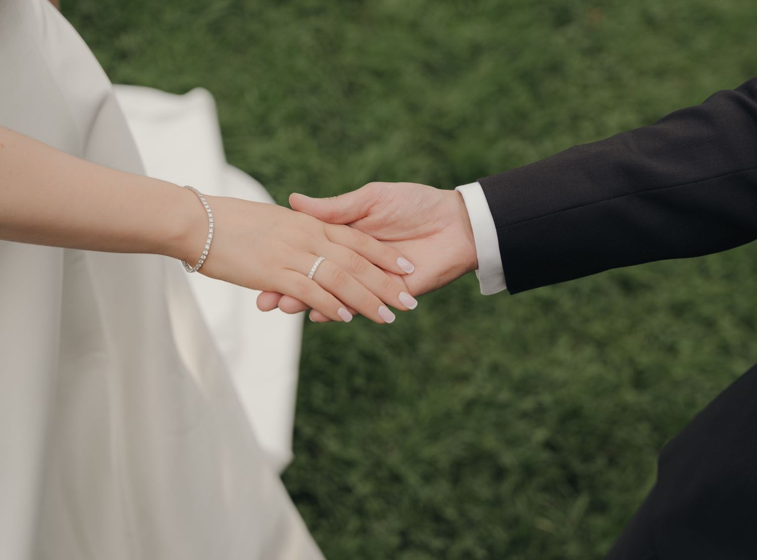 Bride in a white dress and groom in a dark suit hold hands over green grass, her wrist bracelet and ring visible.