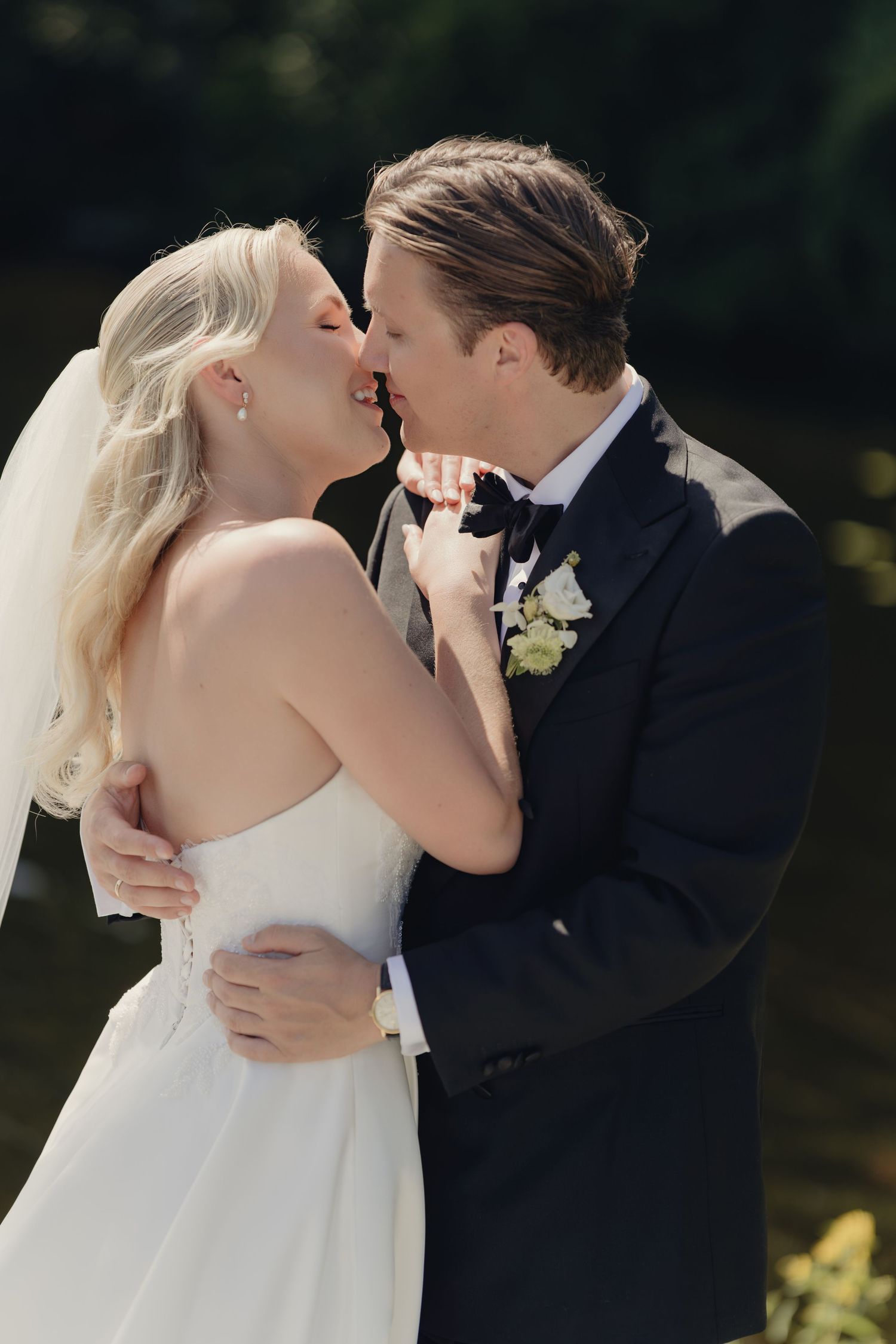 Bride in a strapless white gown and veil shares a kiss with her groom in a navy tuxedo and bow tie outdoors on their wedding day.