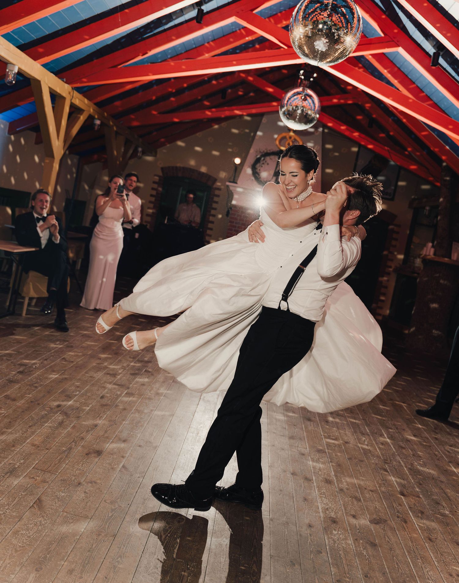 Groom lifting bride in white wedding dress on dance floor under disco ball in decorated barn venue.