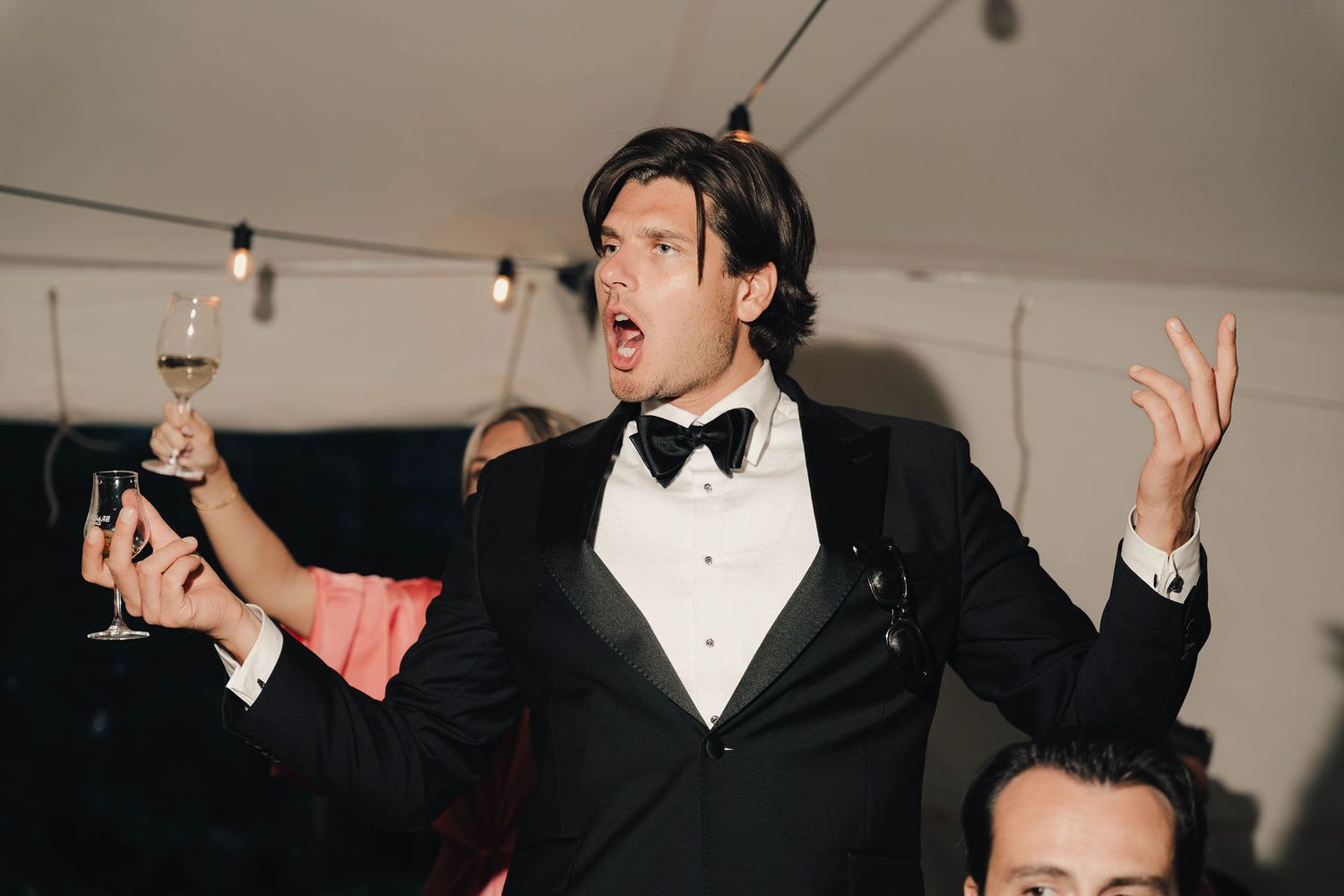 Man in black tuxedo with bow tie holding champagne glass at formal event, looking surprised.