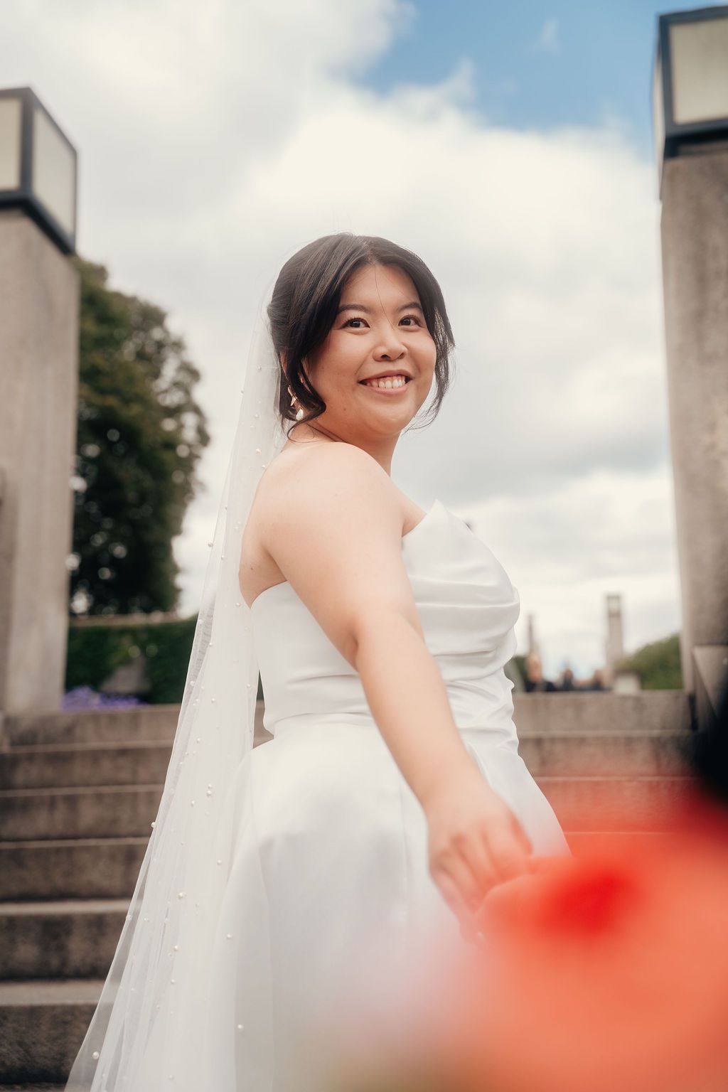 A bride in a white strapless wedding dress smiles while standing on stone steps outdoors on a cloudy day.