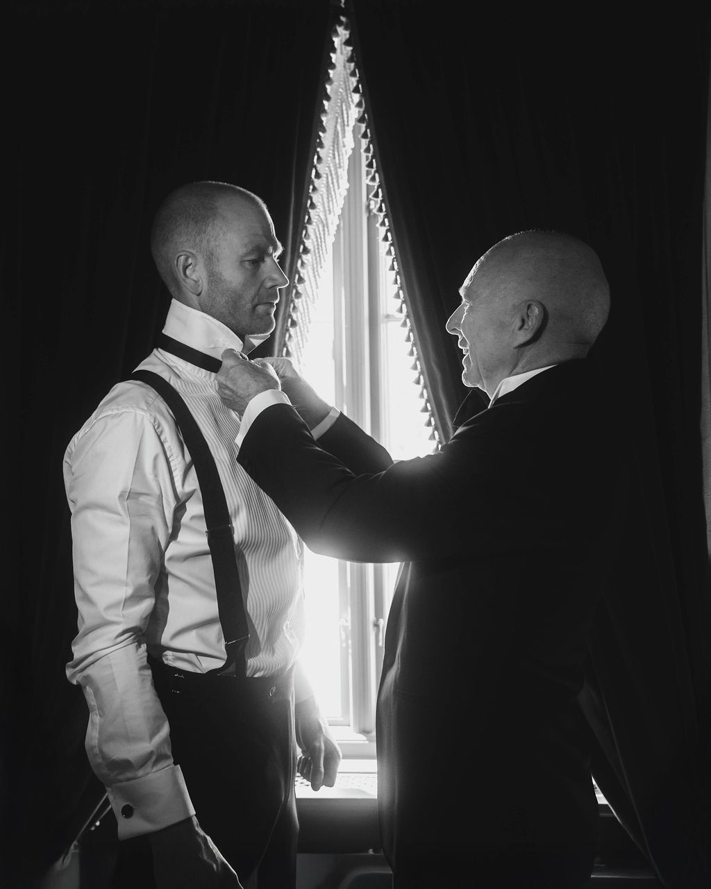 Two figures in white shirts and dark ties stand in silhouette against a bright window in a dramatic black and white scene.