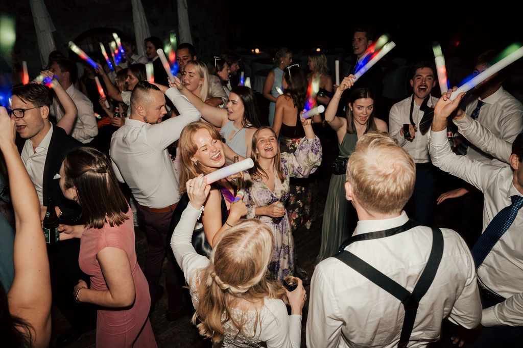 Wedding party guests dance and celebrate with colorful LED glow sticks on a dark dance floor.