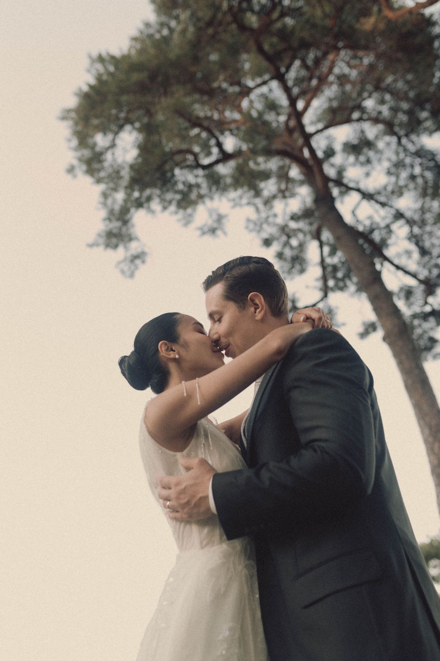 A romantic wedding couple shares a tender kiss under tall pine trees in soft lighting.