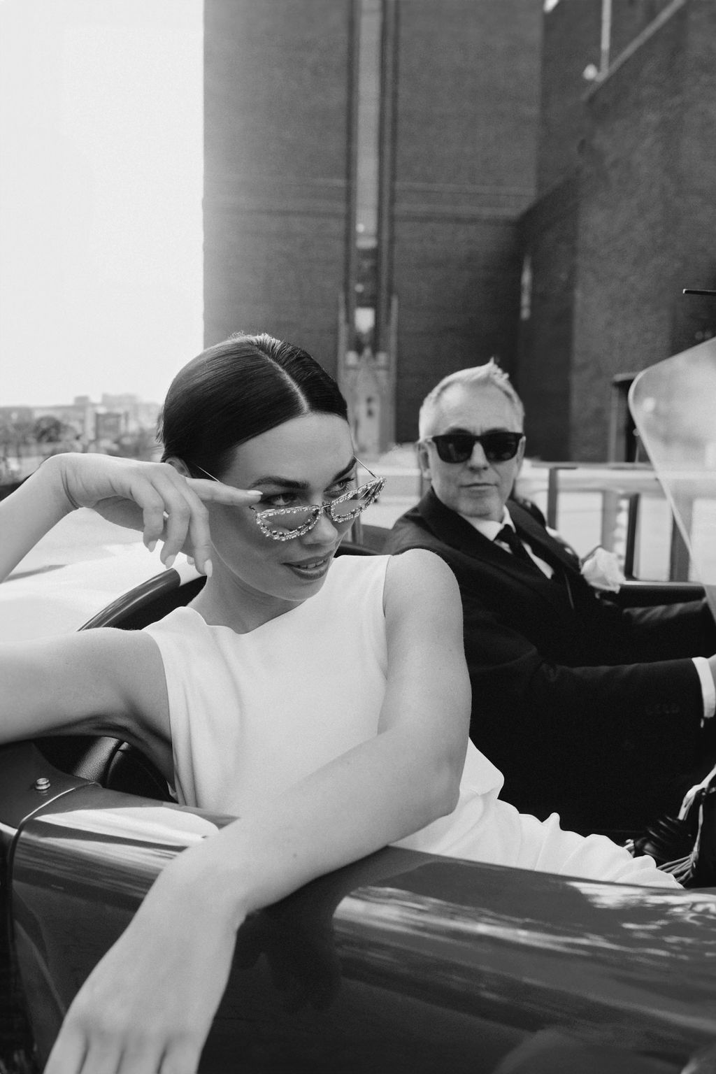 A black and white photo of a person reclining in formal attire against a car door.