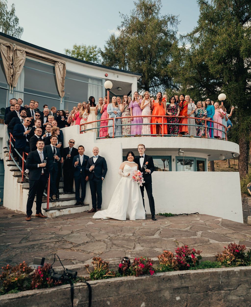 Large group of wedding guests and party members posing together on stone steps outside a modern building.