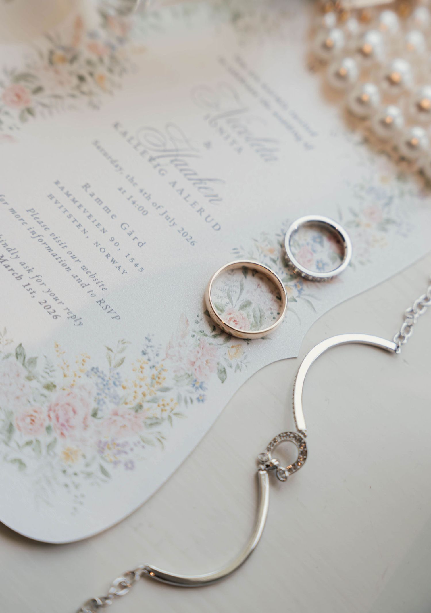 Wedding rings and silver bracelet resting on floral invitation suite with pearl necklace in soft pastel tones.