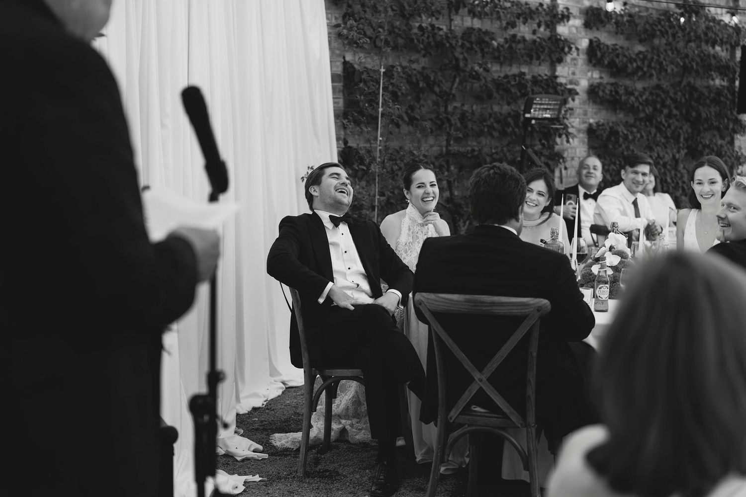 Black and white wedding reception photo of groom laughing at table while guests enjoy speeches.