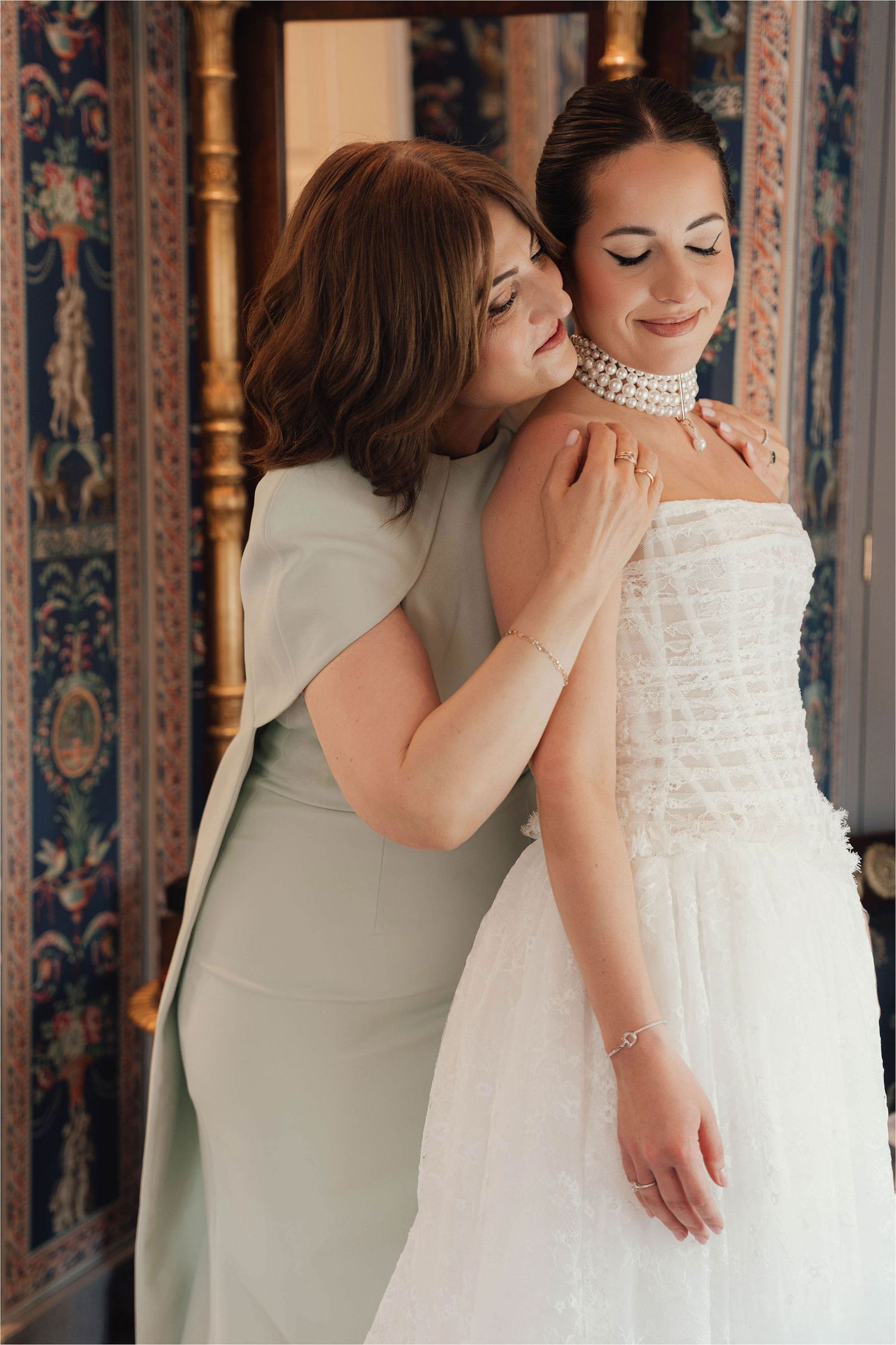 A mother helps her daughter put on a pearl choker necklace as she prepares in her white strapless wedding gown.