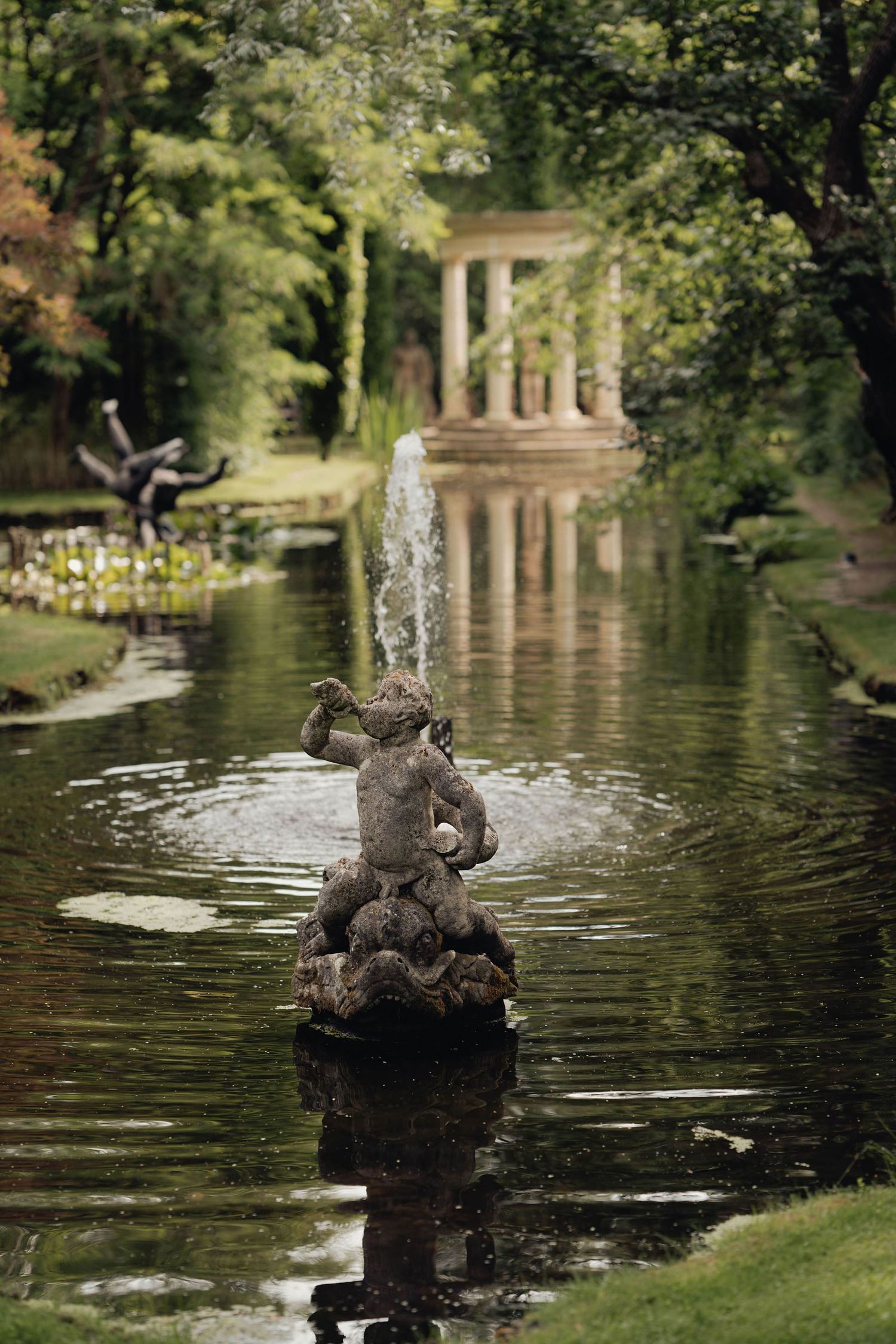 Stone cherub fountain statue in a serene garden pond with a classical columned temple and lush greenery in the background.