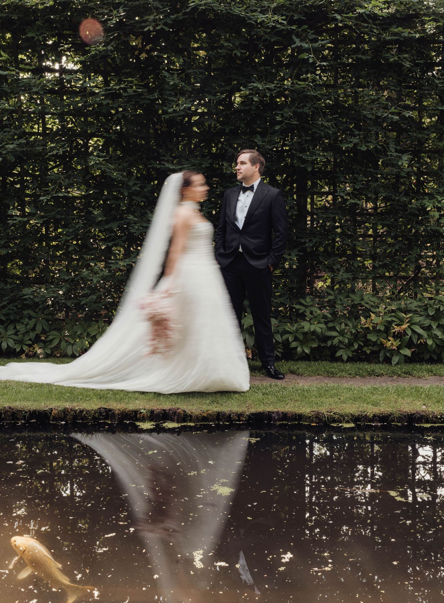 Bride in a flowing white gown and veil stands beside a groom in a black tuxedo by a reflective garden pond with a koi fish.