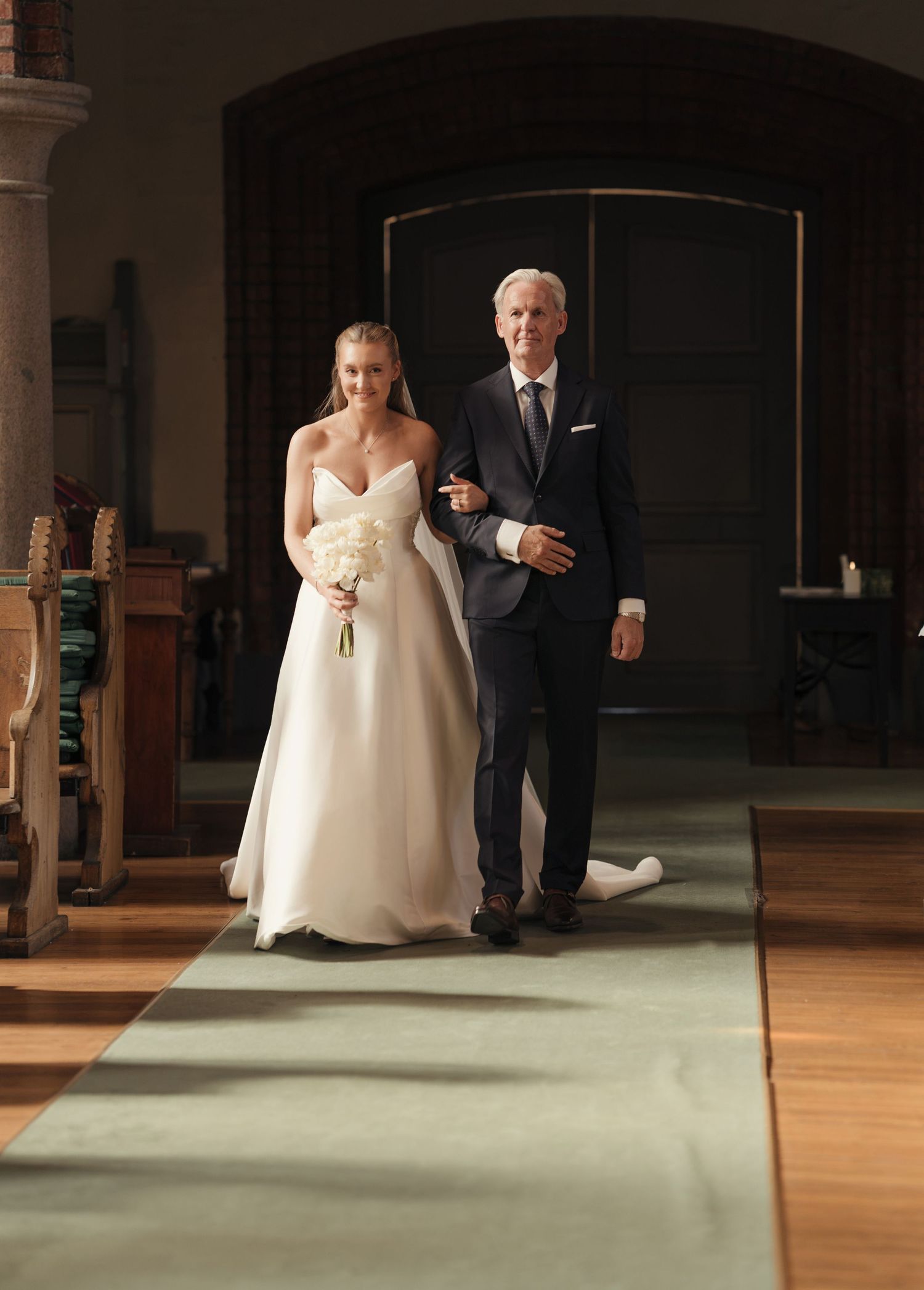 Bride in a strapless white gown holding a bouquet walks arm in arm with a gray-haired man in a dark suit down a church aisle.