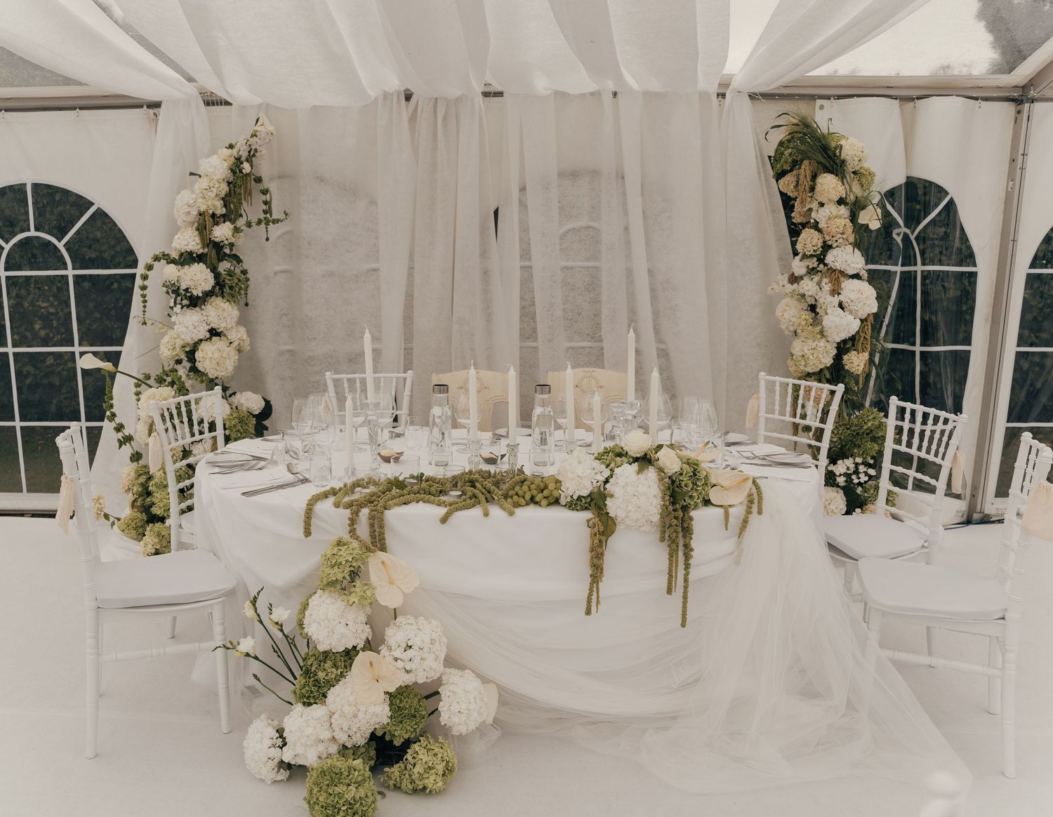 Elegant wedding reception tent with a white draped head table decorated with greenery and white floral arrangements.