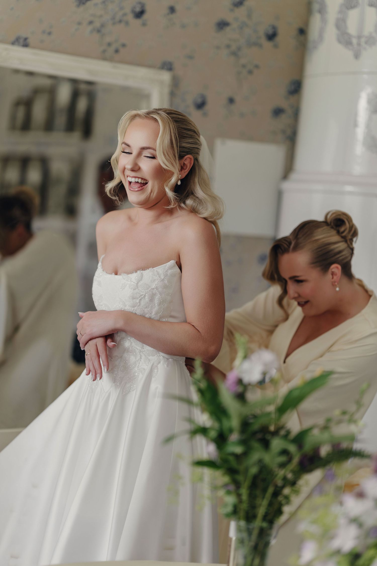 A laughing bride in a strapless white wedding gown stands beside a woman with an updo, with fresh flowers in the foreground.