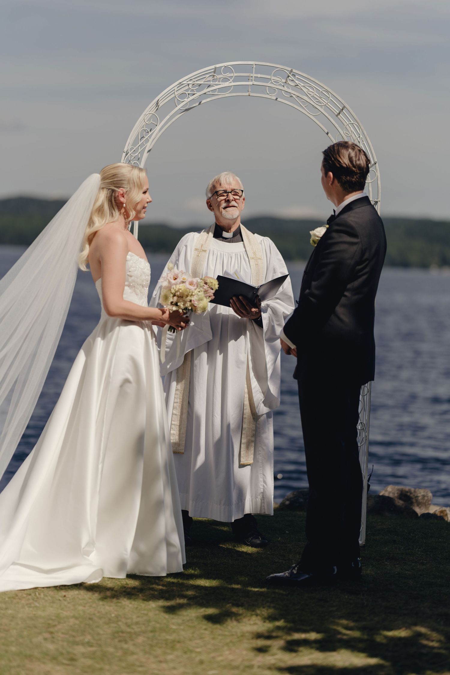 Bride in a satin gown and long veil faces the groom as an officiant leads their lakeside wedding ceremony.