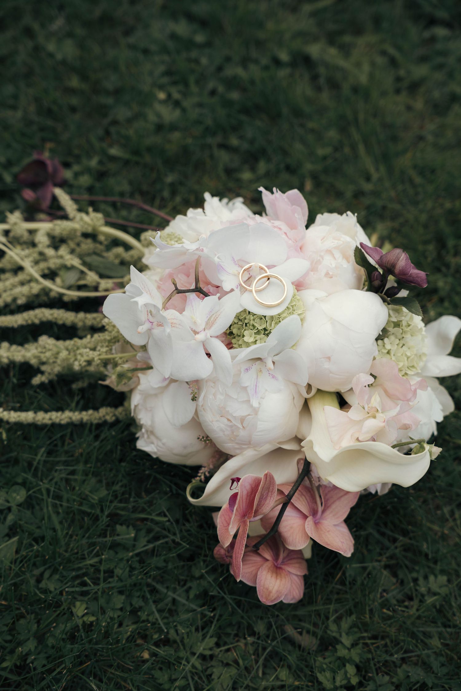 Wedding bouquet of white peonies and pink hydrangeas with gold rings on green grass.