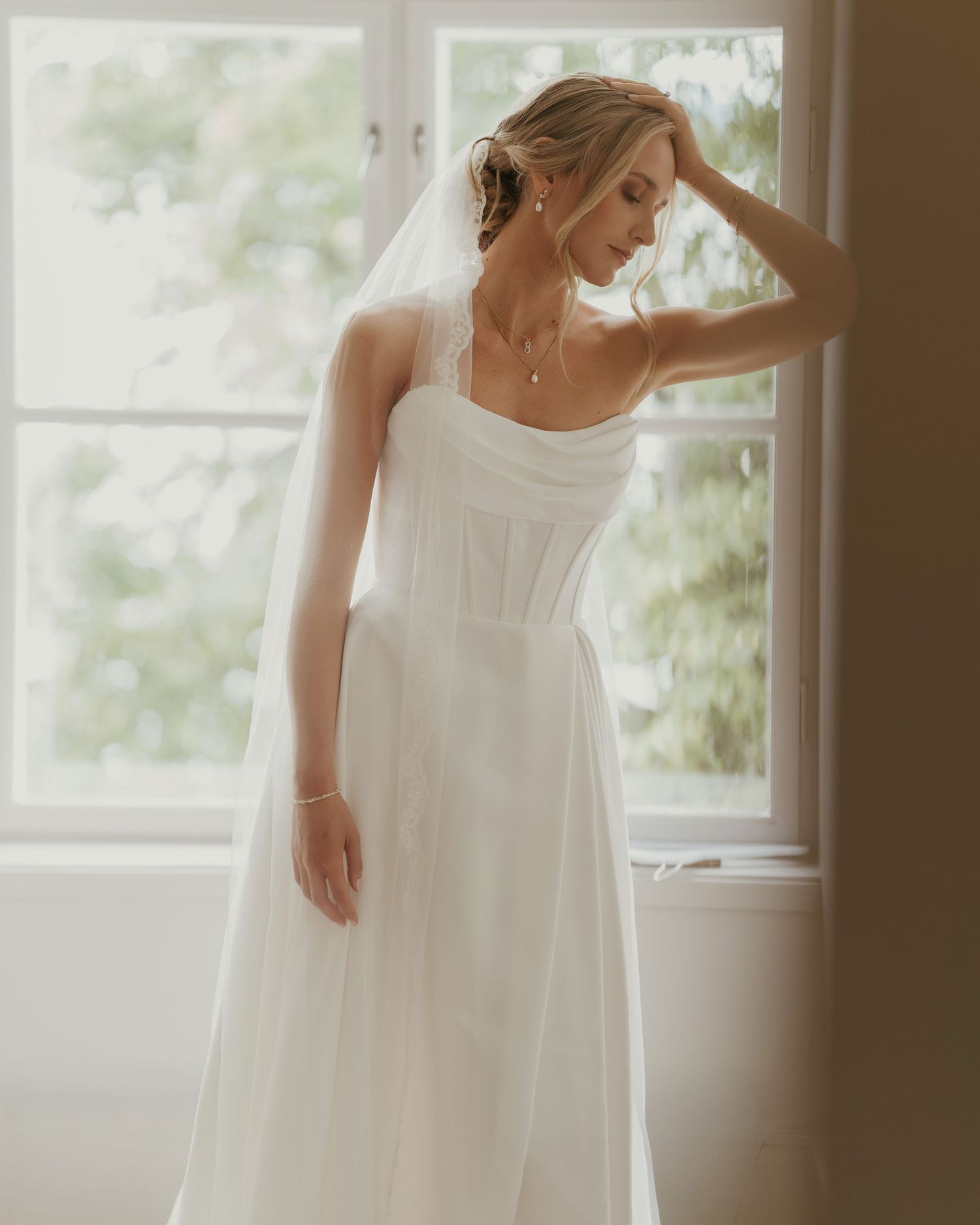Bride in a white A-line wedding gown with corset bodice and veil, posing by a sunlit window.