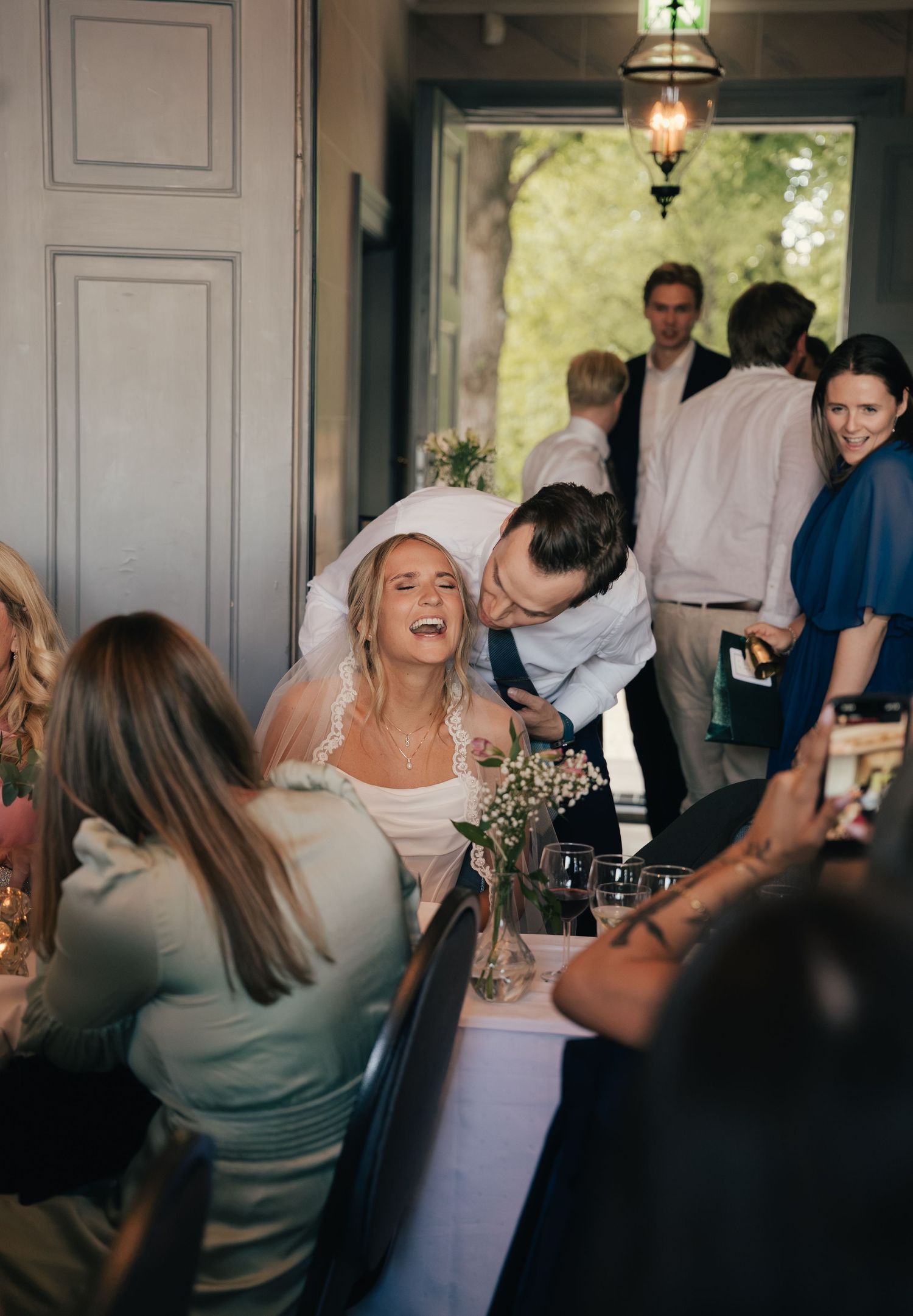 Laughing bride in veil seated at reception table as groom leans in to whisper at indoor wedding dinner.