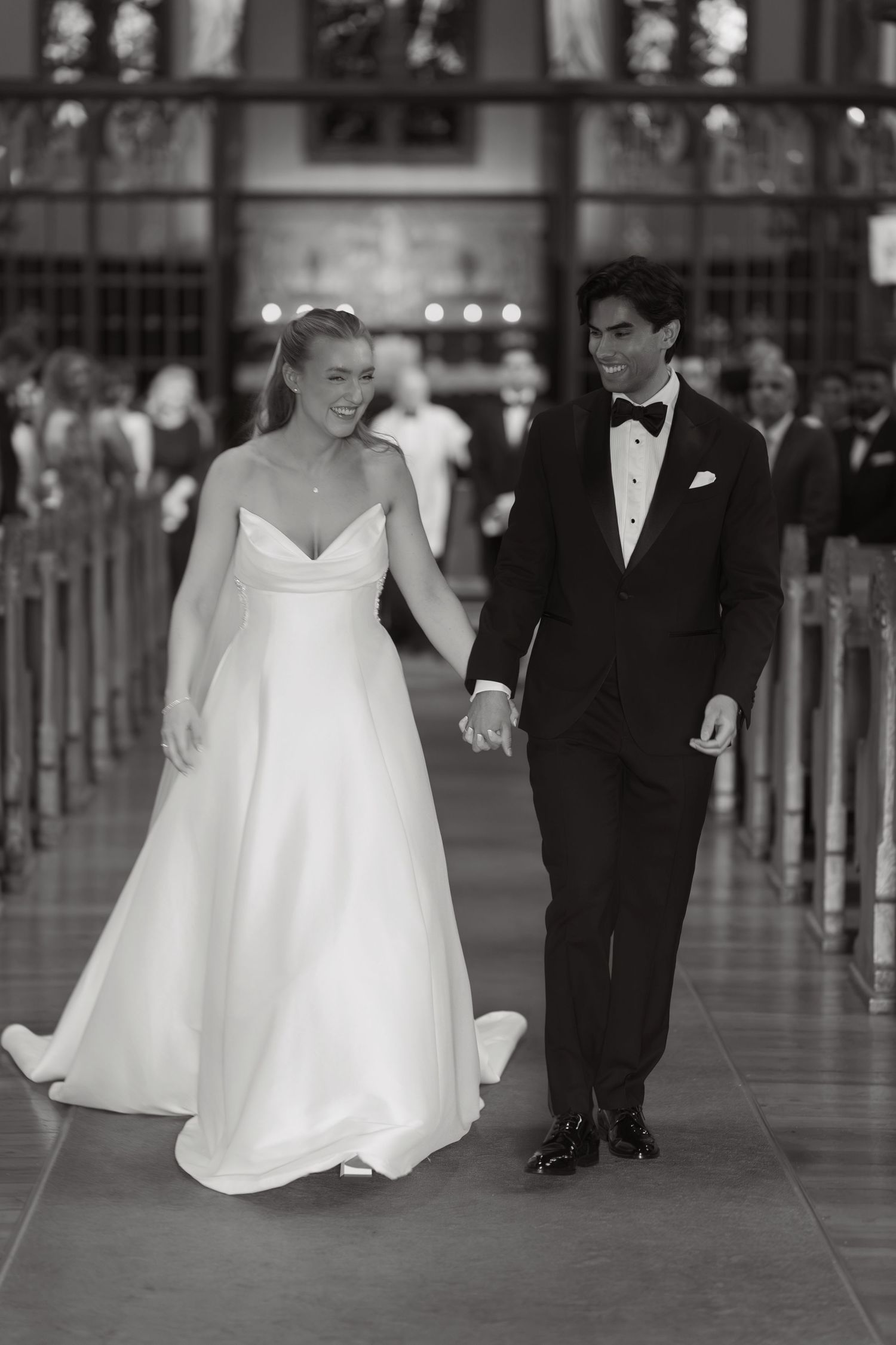 A smiling bride in a flowing white gown holds hands with her groom in a tuxedo as they walk down the church aisle in black and white.