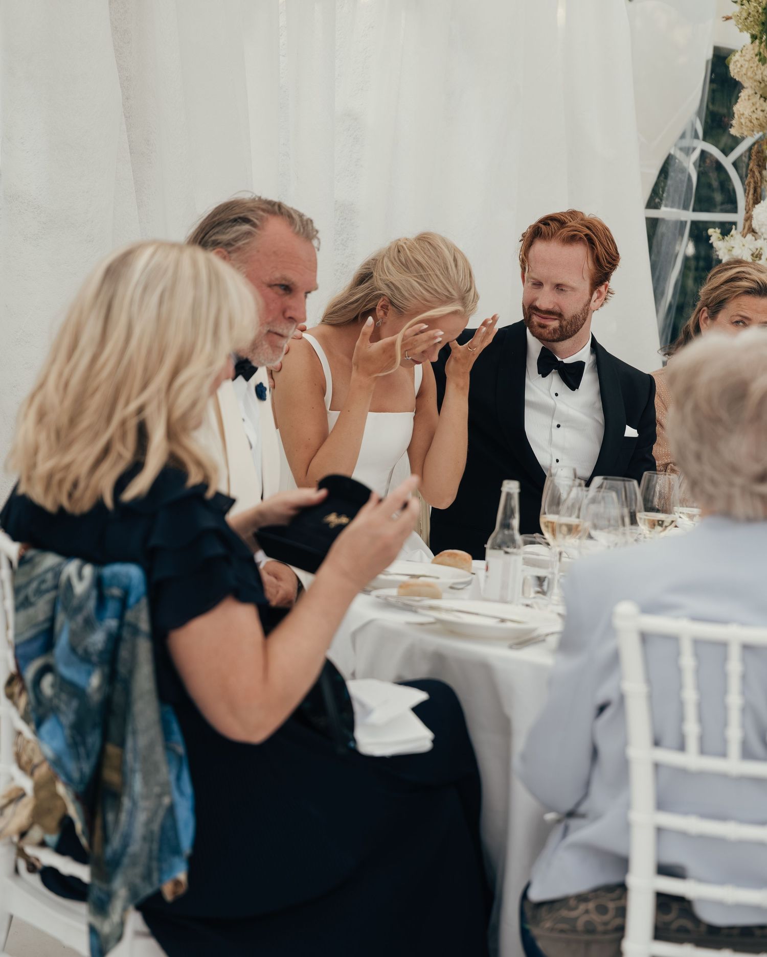 Wedding guests in formal attire sit at a white-clothed reception table, laughing and wiping away tears during the celebration.
