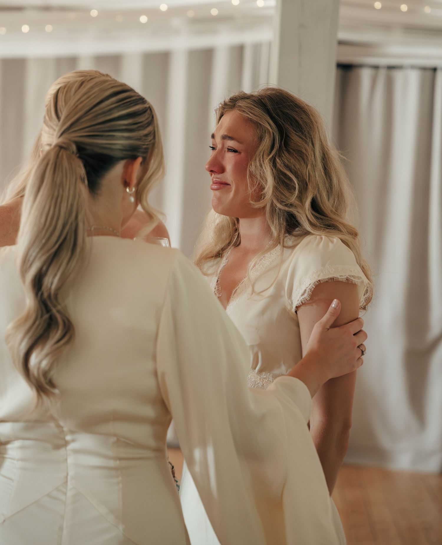 Teary-eyed woman in a cream dress is comforted by the bride in a white gown with a ponytail during a wedding reception.