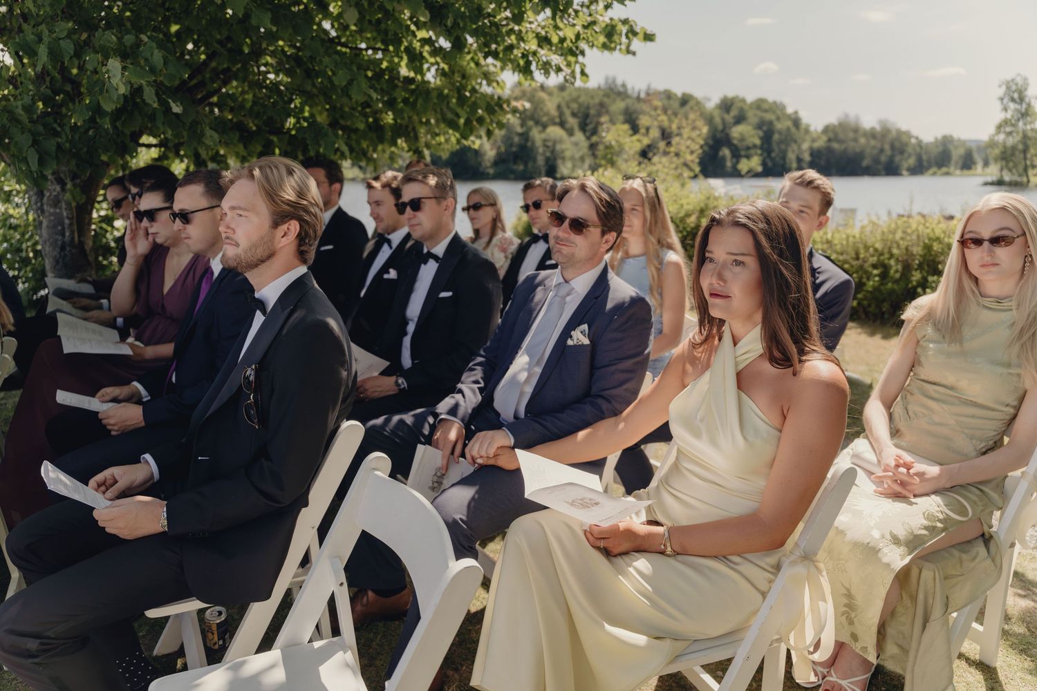 Wedding guests in formal attire sit on white folding chairs during an outdoor lakeside ceremony surrounded by trees.