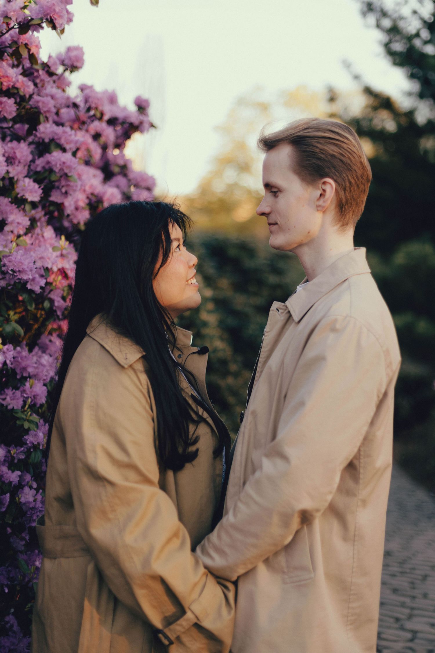 Two people in beige coats share an intimate moment beneath blooming pink cherry blossoms in a park setting.