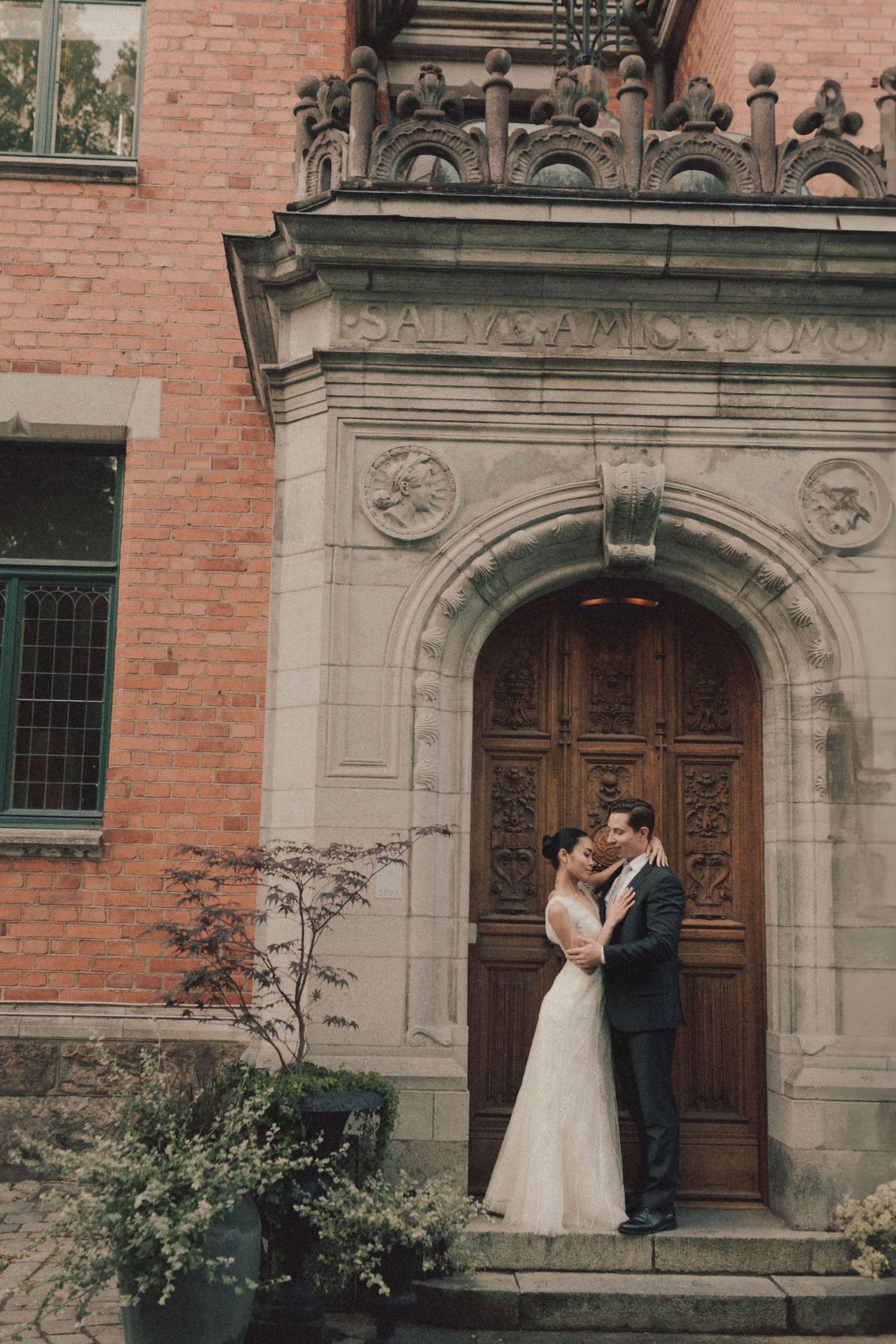A romantic couple shares a tender moment in front of an ornate stone doorway of a historic brick building.