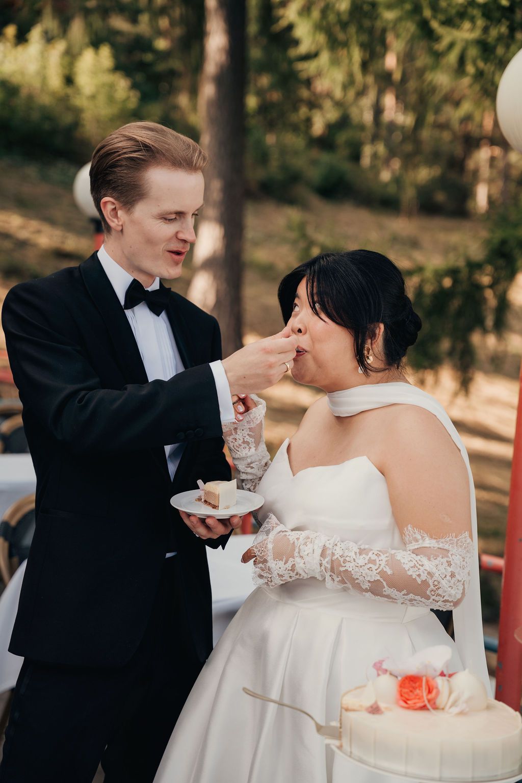 A couple in wedding attire shares a playful moment while cutting their white wedding cake outdoors.