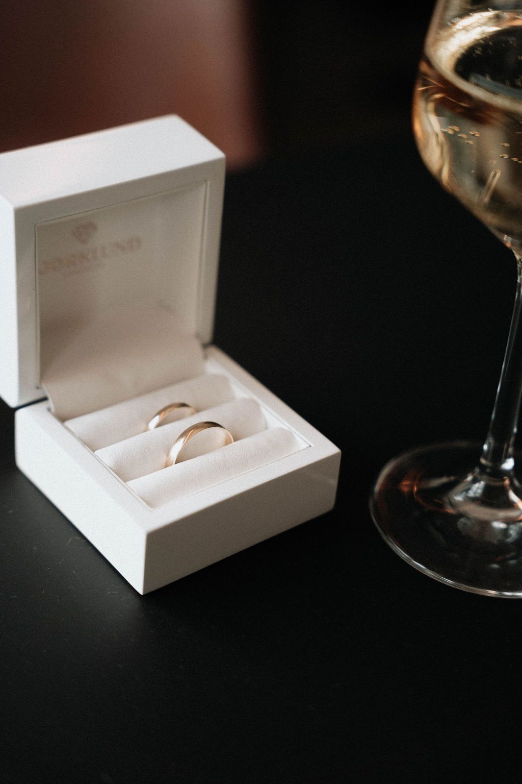 A white wedding ring box with two gold bands sits beside a glass of white wine on a dark surface.