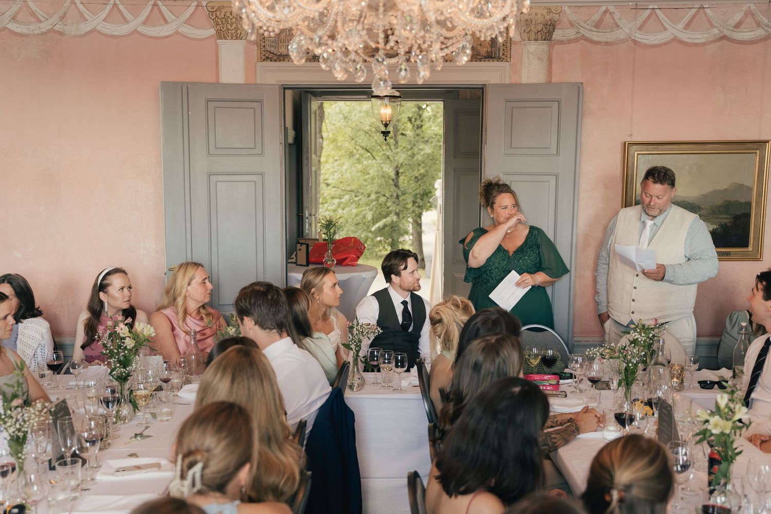 Wedding guests seated at elegant pink dining room table during reception speech under crystal chandelier.