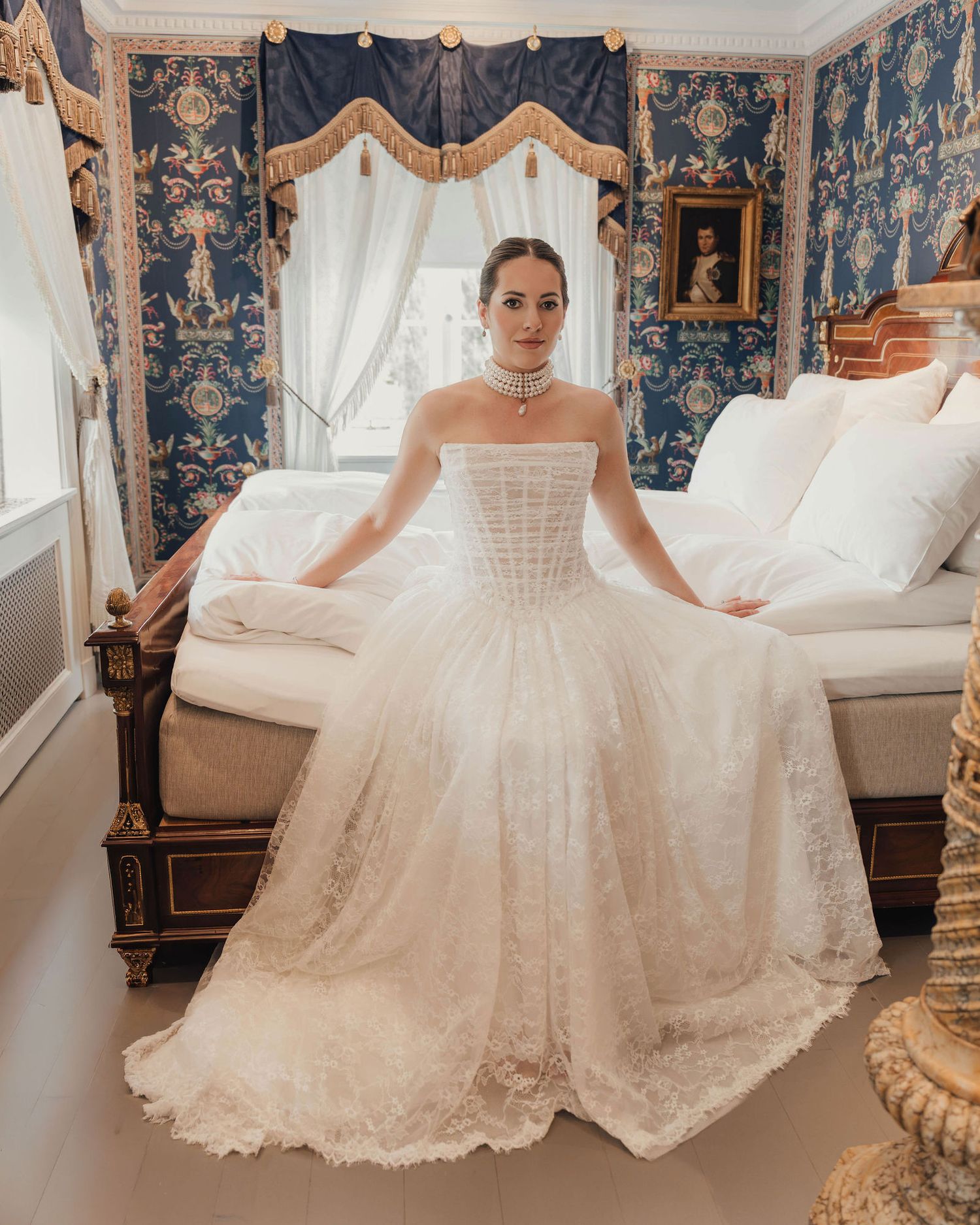 Bride in strapless lace ball gown wedding dress posing in ornate blue floral wallpapered bedroom.