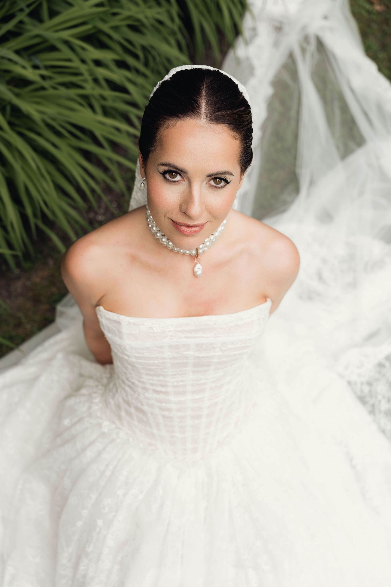 Elegant bride in strapless white ball gown with diamond necklace and earrings, wearing a veil outdoors.