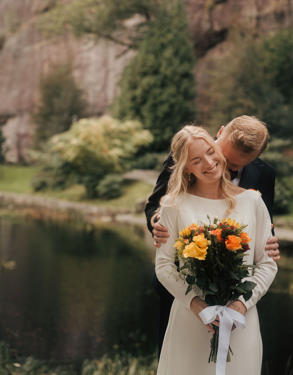 A couple shares romantic moments by a lake with sunflowers and mountains in the background during their wedding day.