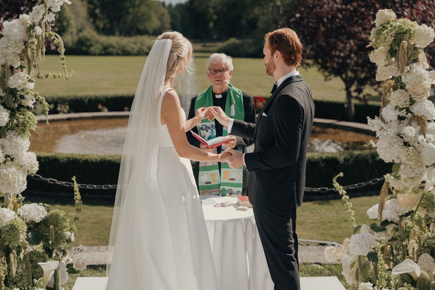 Bride in a long veil and groom in a dark suit exchange vows before an officiant at an outdoor garden wedding framed by white floral arrangem