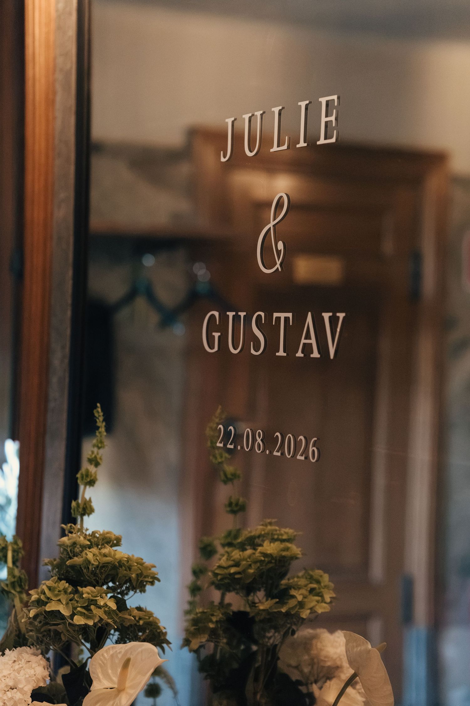 Mirror with the names Julie & Gustav and the date 22.08.2026 written in elegant script, reflecting green and white wedding flowers.