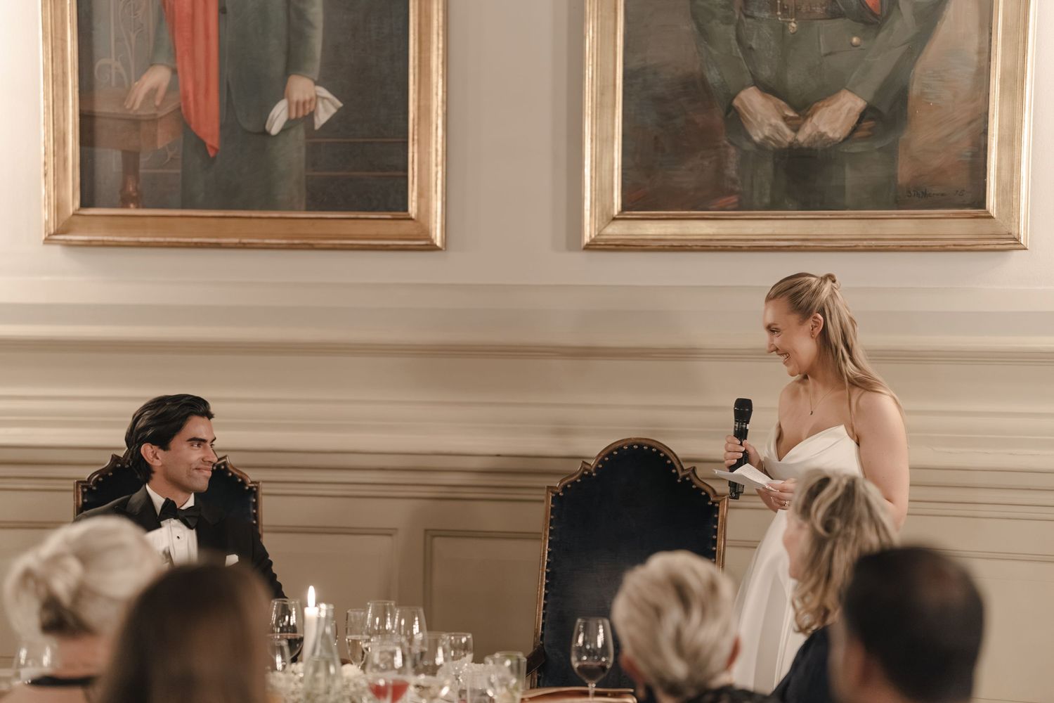 Bride in a white gown holds a microphone and gives a speech to a seated groom beneath two framed portraits at a formal dinner.